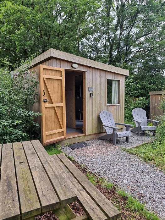 a wooden cabin with an open door, a window next to a picnic table and two outdoor chairs