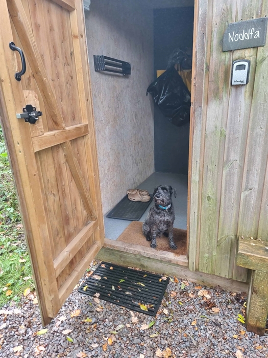A small black dog sits on a mat in the doorway of a wooden cabin