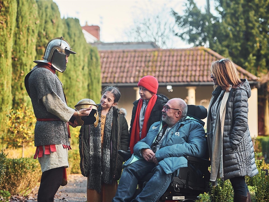 A man dressed as a Roman Gladiator talks to a family in the museum garden.