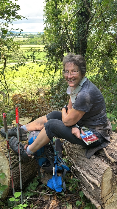 Smiling hiker resting on a fallen tree in woodland, wearing walking boots and glasses, with trekking poles and a backpack beside her. Green countrysid