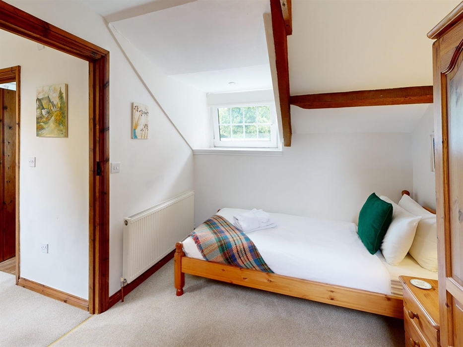 A bright twin bedroom with sloping ceiling, wooden furniture, tartan throw, and a window overlooking greenery.