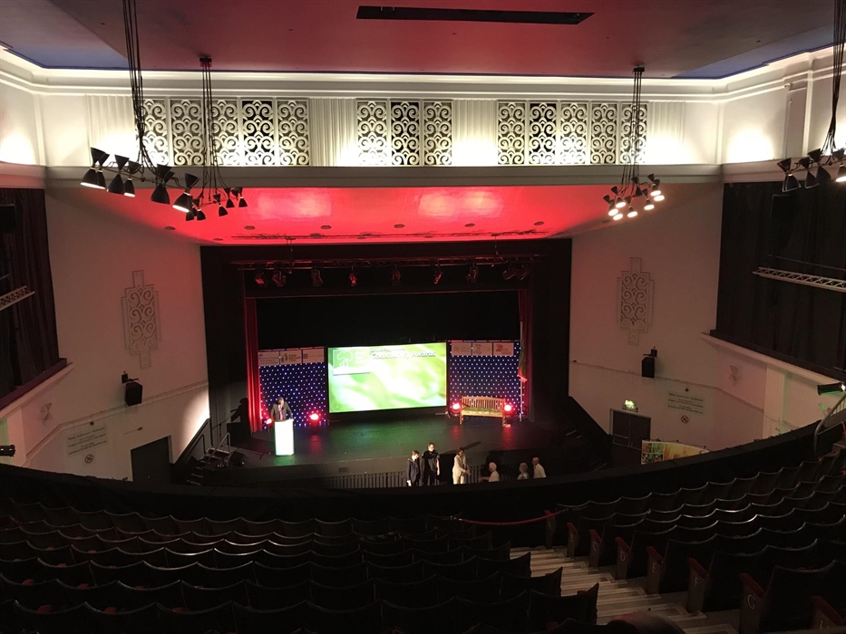 Looking down from the Balcony, over the fixed seating, to the stage below, lit in red with large central presentation screen and lectern to the left