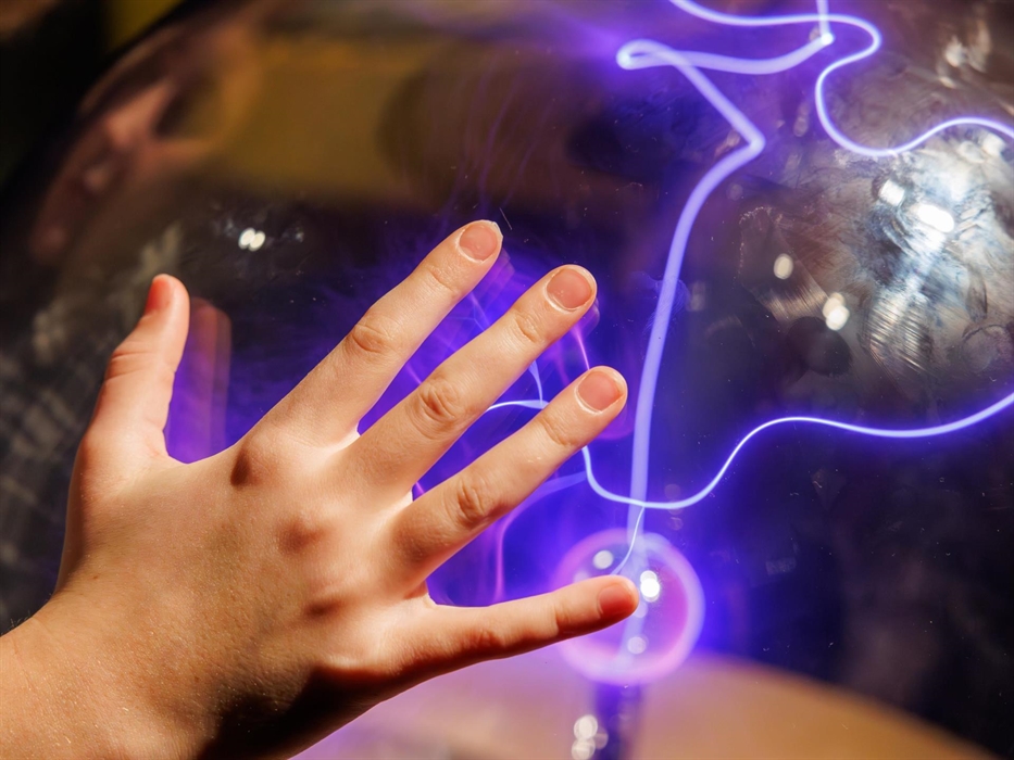 A child resting their hand on the case of the Plasma Sphere exhibit, attracting coils of electricity to their fingertips.