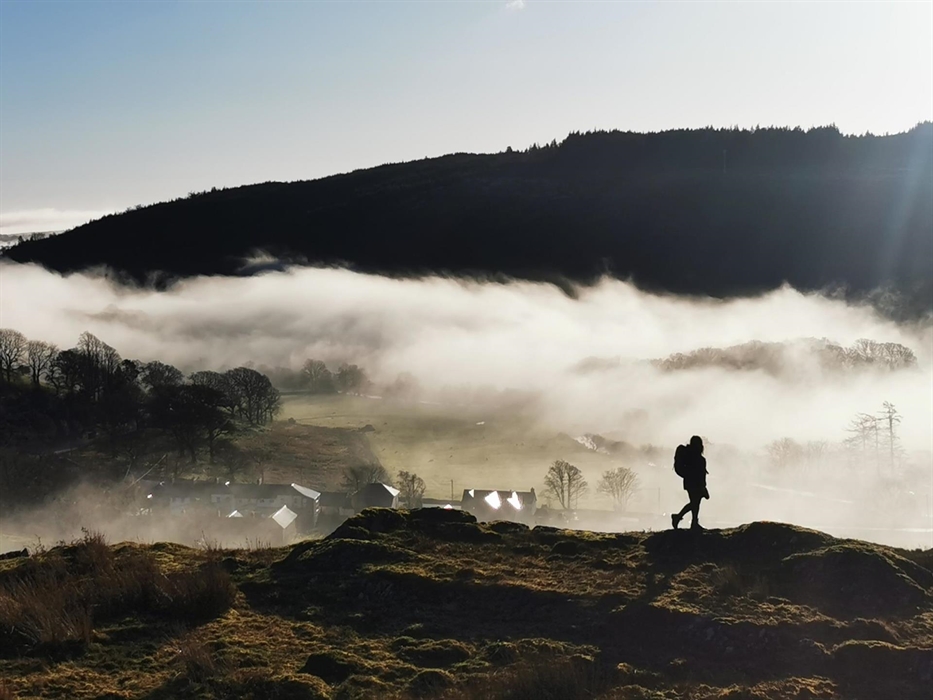 The silhouette of a woman wearing a backpack standing on a hill with a background of mist and trees in morning sunlight