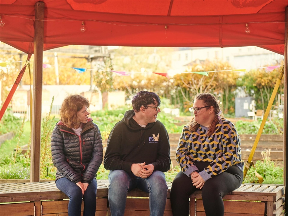 three young people are chatting together, outside in the museum's garden. They're sitting on a wooden bench under an orange canopy.