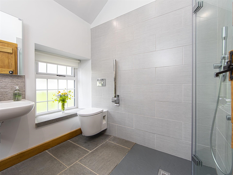 A modern bathroom featuring a walk-in rainfall shower, wall-mounted toilet, large frosted window with flowers, and light grey tiles with natural slate