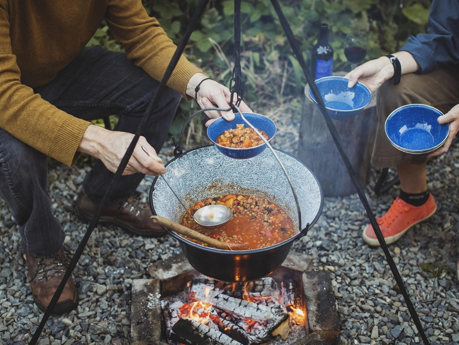 A campfire with people surrounding it. A large pot hangs above full of a steaming stew which is being ladled out into bowls.