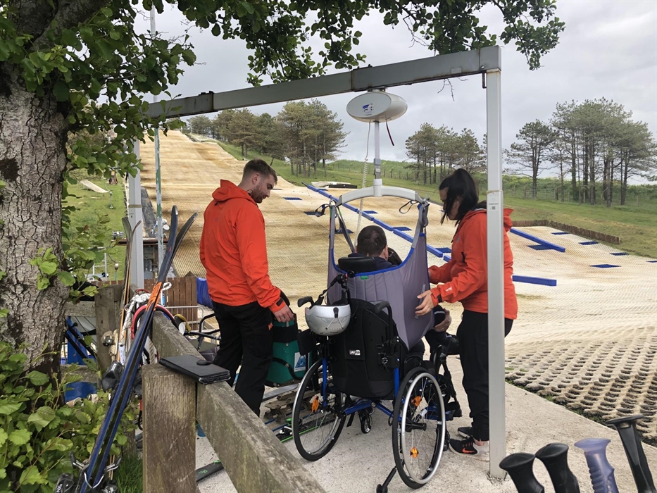 Two volunteers in orange jackets help a man in a grey sling be hoist transferred from wheelchair to sit ski.