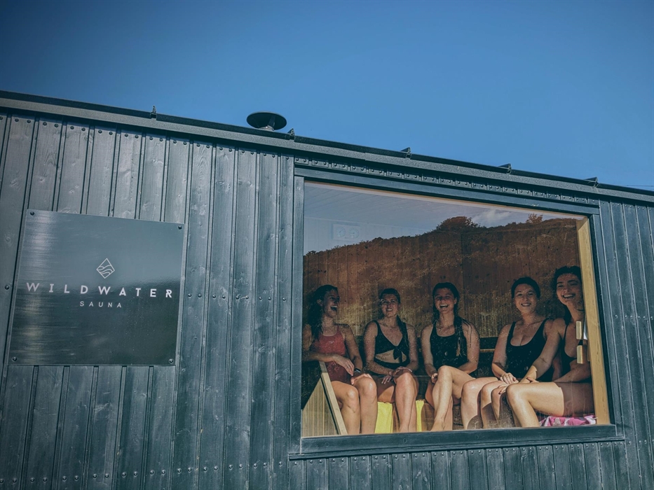 Five happy ladies sat in a wooden sauna looking out of a panoramic window towards the sea