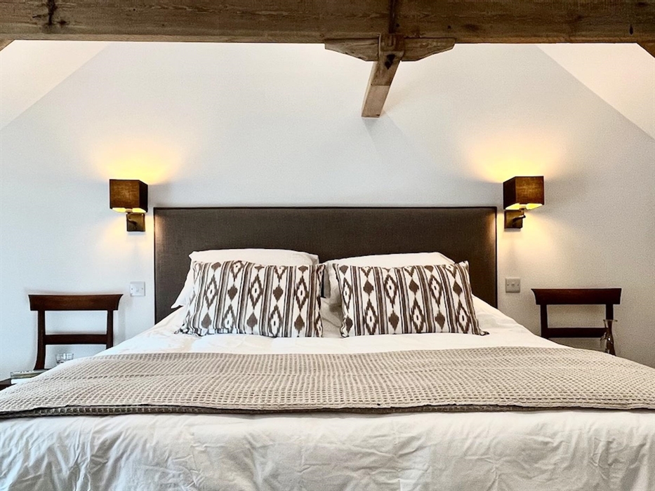 A tasteful neutral coloured bedroom with exposed beams and large bed with decorative cushions and throw, with an antique chair to each side of the bed