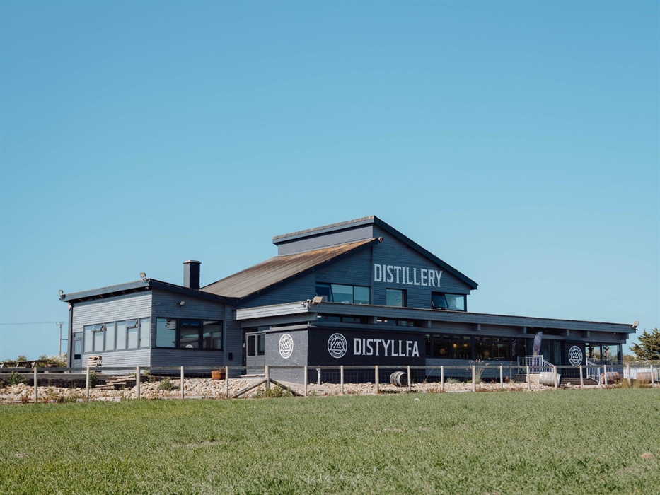 A picture of In the Welsh Wind Distillery - grey bilding with white 'distillery' 'dystyllfa' wording in front of a field of green crop
