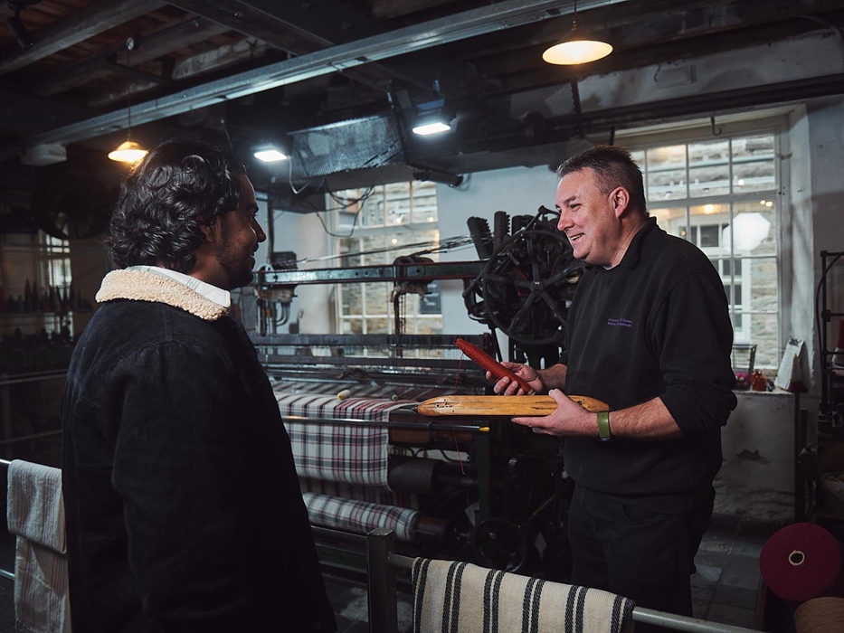 A craftsman explains the weaving process to a visitor. He holds a bobbin of red thread and a wooden shuttle, which passes through the loom to weave th