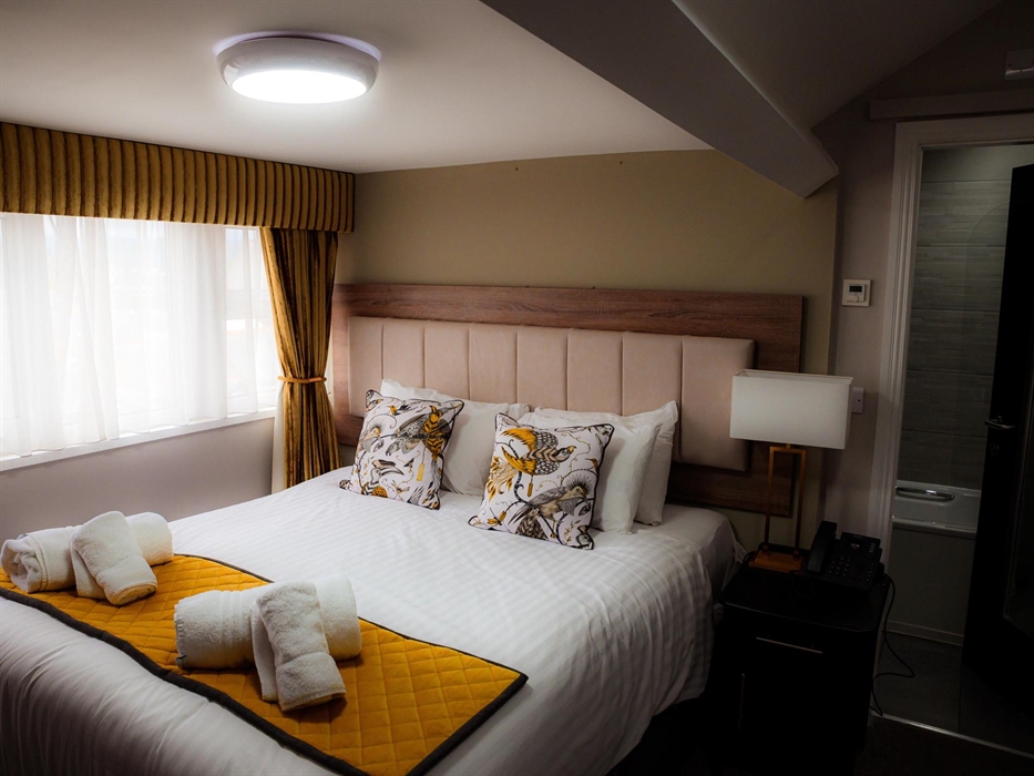 A stylish, inviting hotel room with a plush double bed, elegant patterned pillows, warm lighting, and a modern en-suite bathroom.