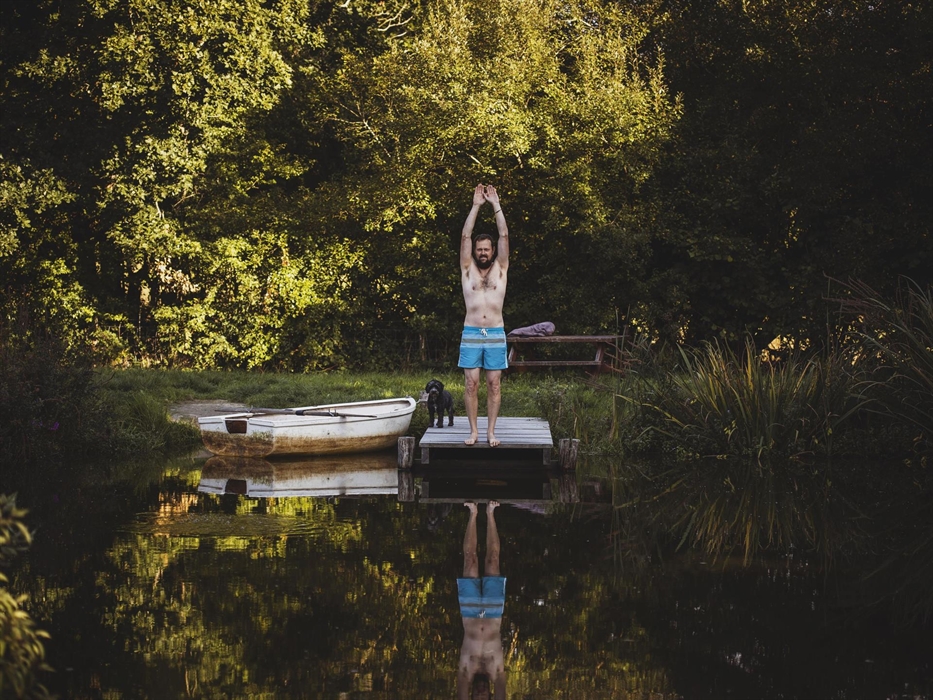 A man standing on a jetty over some water ready to dive. A rowing boat sits to the side of the image. The landscape is woodland.
