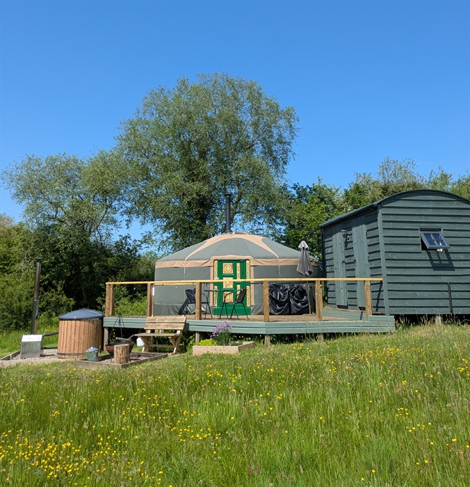 A dark green and beige yurt  with a green and pink painted door and a dark green wood hut on a raised deck with a wood hot tub to one side and a fire