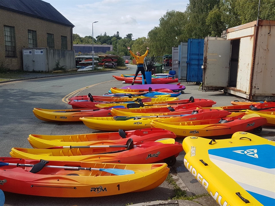 Lots of colourful kayaks and paddleboards ready to be issued to customers with an excited person jumping in the air