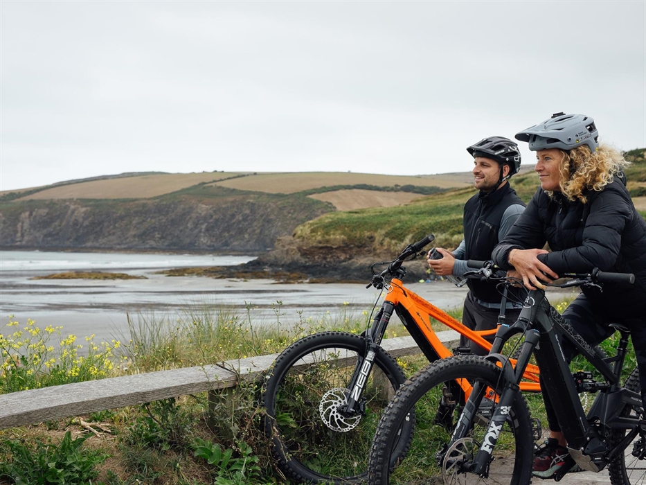 Two cyclists with electric mountain bikes look out across Newport beach.