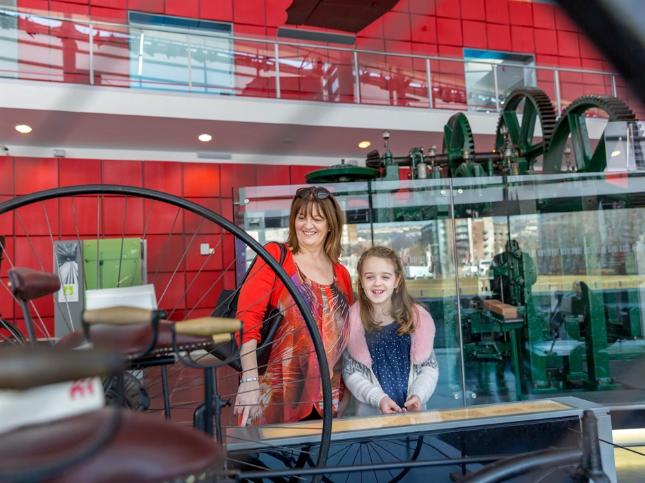 A mum stands with her young daughter inside the museum. They are looking at an early bicycle on display. The mum wears a red patterned dress and the g
