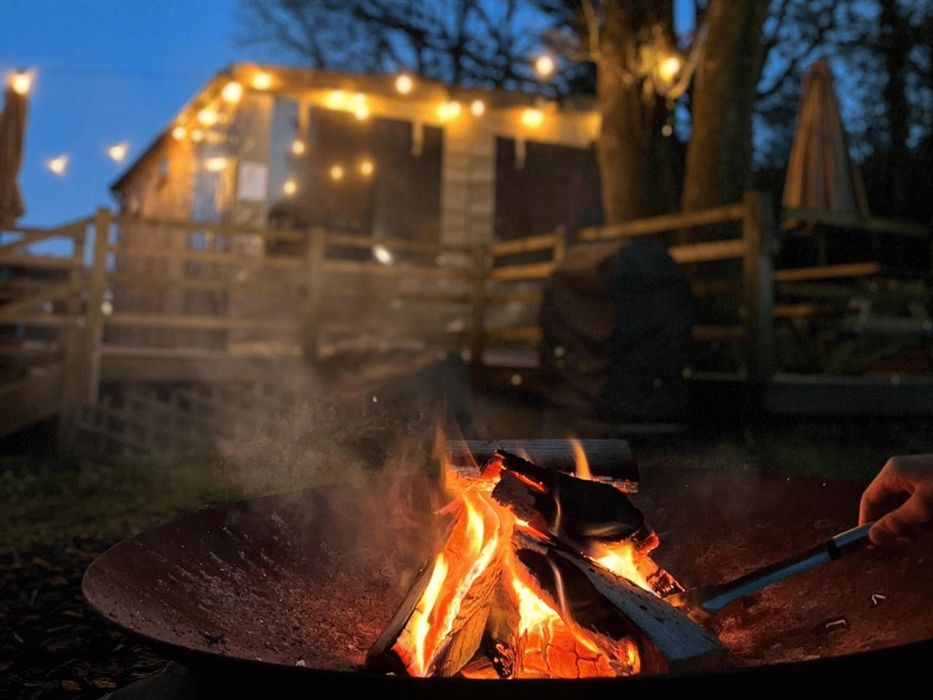 Its night time and a small fire is burning in a fire bowl with a hand stoking the logs. In the background is a wooden cabin lit up by string lights