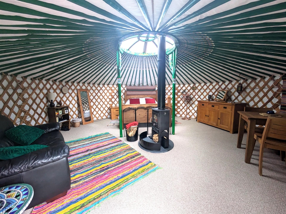 The yurt interior is round with many green painted rafters. There is wood bed at the back, wood table, chairs and cupboard to the right, a black leath