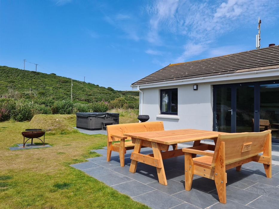 Outside wooden seating set on a slate patio, six seater hot tub, firepit and BBQ visible on grass in front of the corner of Ty'r Felin a self catering