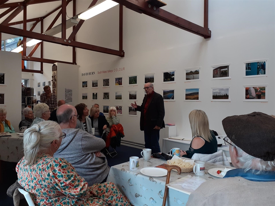 figure standing in a gallery space interior surrounded by other figures seated around tables  listening to them. On the wall behind the standing figur