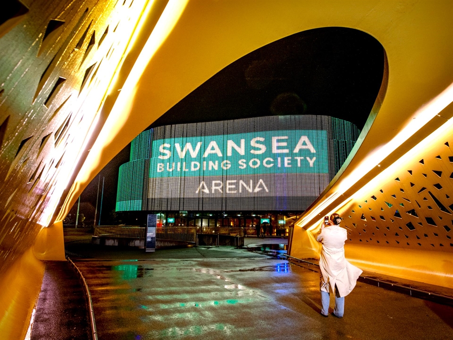 An image of the Swansea Building Society Arena logo projected on the side of the building, taken from the a gold bridge. There is a person in a long c
