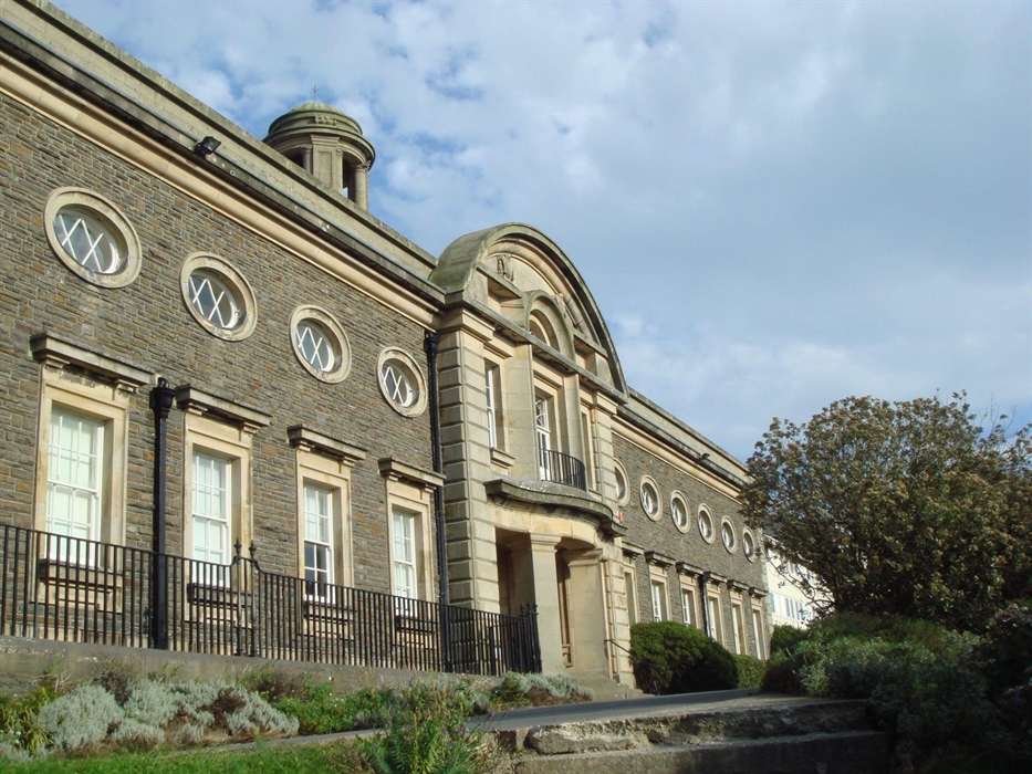 Edwardian building with a sandstone porch with columns and oval windows above large square windows on either side of the entrance. Bushes and an iron-