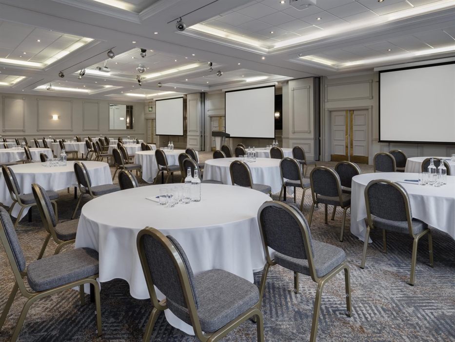 A large spacious meeting room with round tables, mounted screens and projectors.