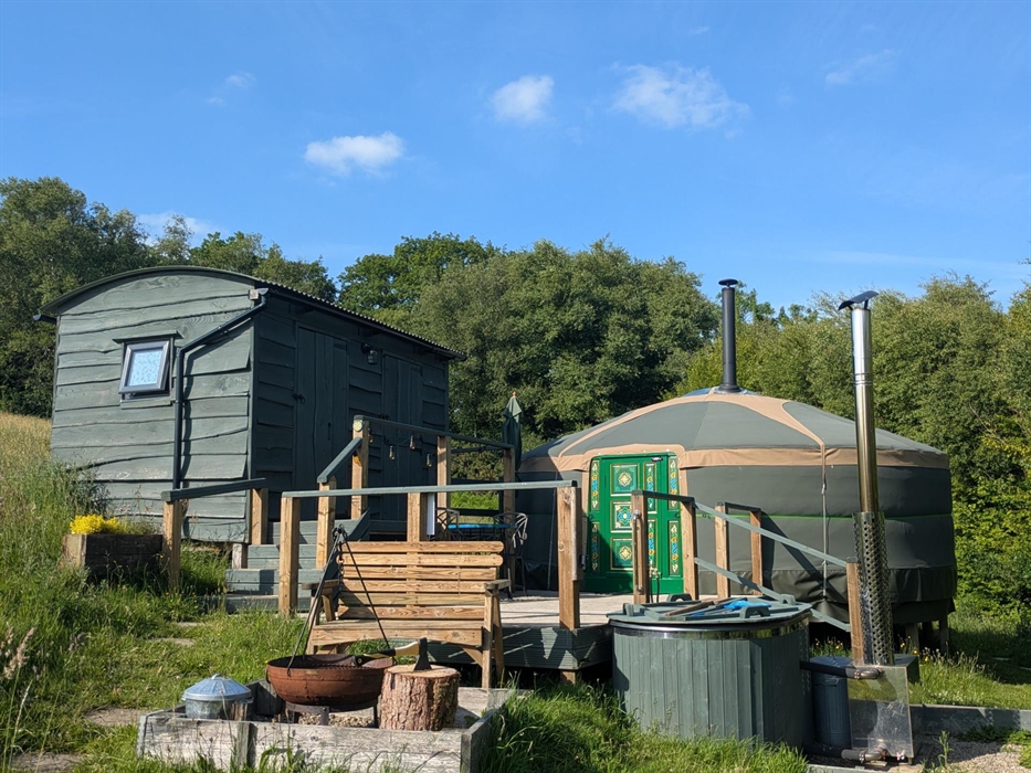 A green yurt with a bright green decorated door, on a wood deck with a dark green painted wood hut up some steps on the left of the image, a dark gree