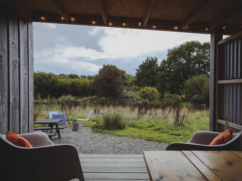 View from inside an open camp kitchen of a campfire scene and trees and hills in the background.