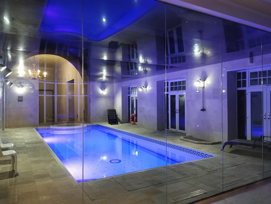 Elegant indoor swimming pool at Plas Cilybebyll, illuminated by blue mood lighting and surrounded by stone walls, lounge chairs, and arched doorways.