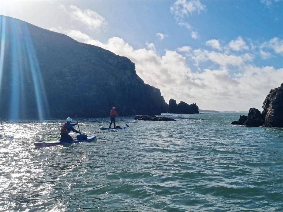 Two people paddleboarding on the sea next to cliffs, on a cold sunny day