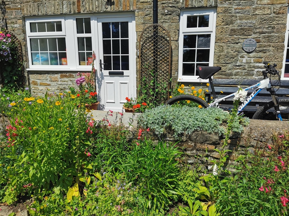 Close-up of a stone cottage entrance with a white door, colourful flower garden, and a bicycle resting against a low stone wall.