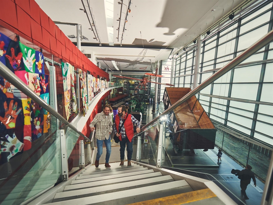 two men are walking up an open stair case inside a light, modern gallery. Behind them you can see a large coal truck that would have been pulled by tr