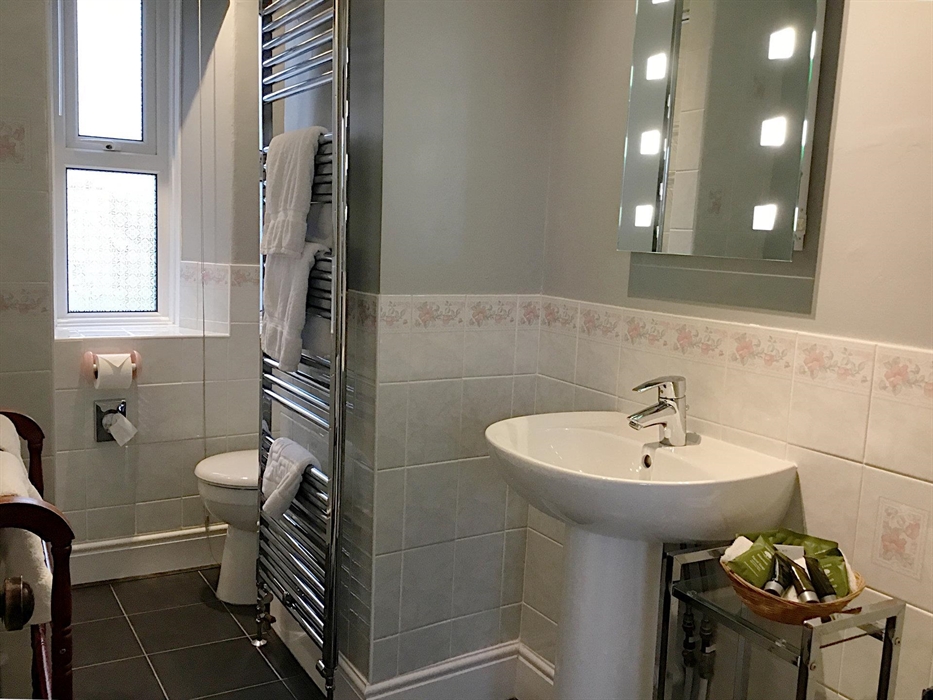 Twin room en-suite showing basin with a lit mirror above, and complementary toiletries and white towels on a towel rail radiator