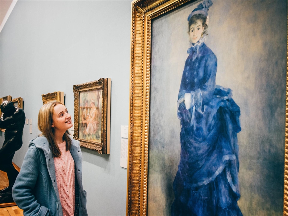 young lady looking at painting by Renoir called La Parisienne. Painting shows a young woman wearing a blue dress.