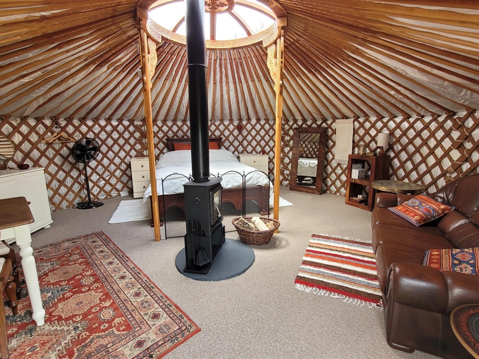 Yurt interior is round, with many carved wood rafters radiating from a carved wood central roof light. Wood bed to rear, brown leather sofa to right,