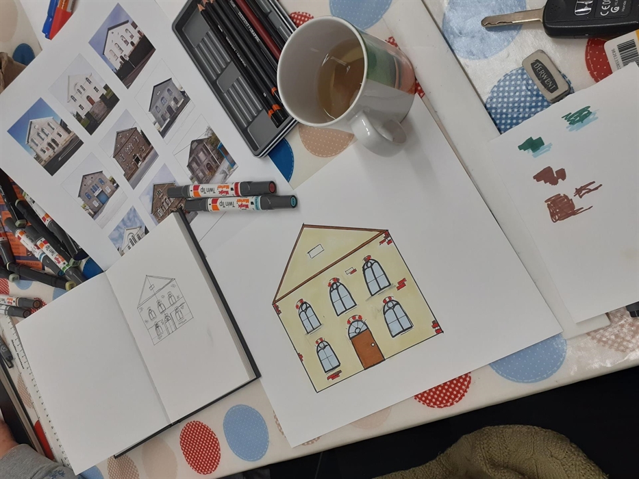 Table with drawing activity happening on it. Table has spotty tablecloth upon which there is a poster of chapel photographs by Paul Cabuts, an open sm