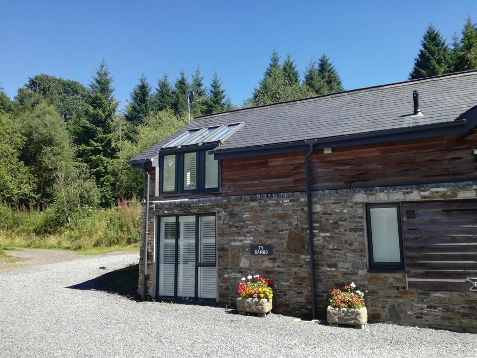 A converted stone barn nestled at the forest’s edge, featuring solar panels, shuttered windows, and vibrant flower displays by the entrance.