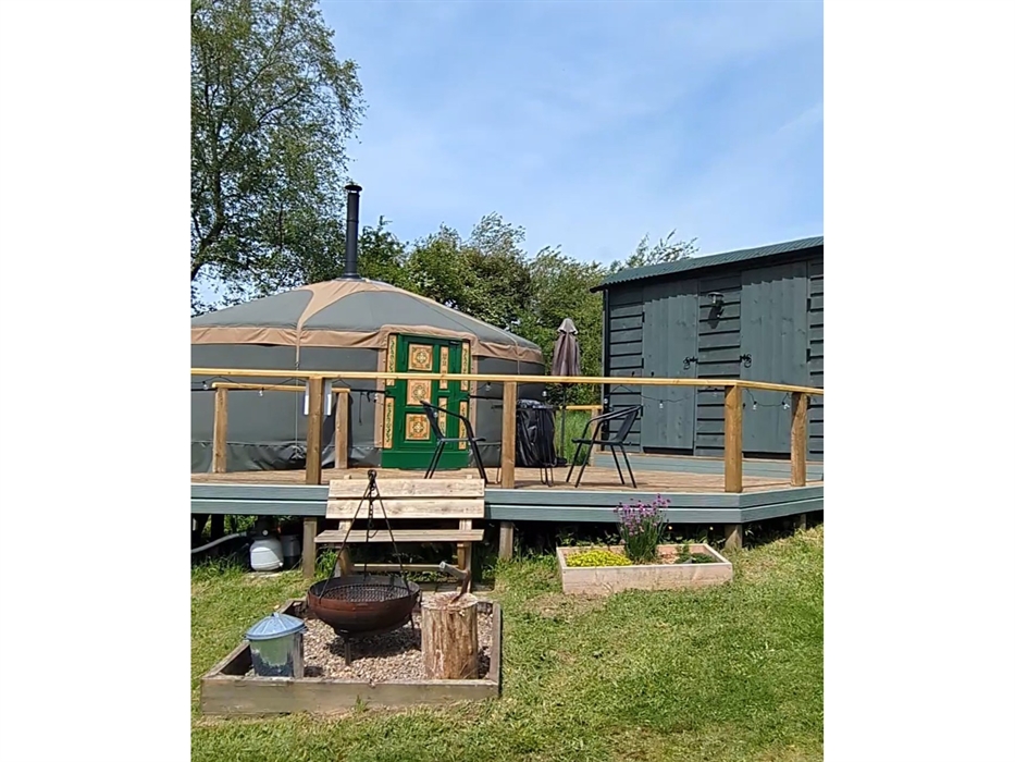 a steel fire pit with tripod in a pebbled enclosure in front of a wood bench, which is in front of a raised deck with a yurt and hut off.