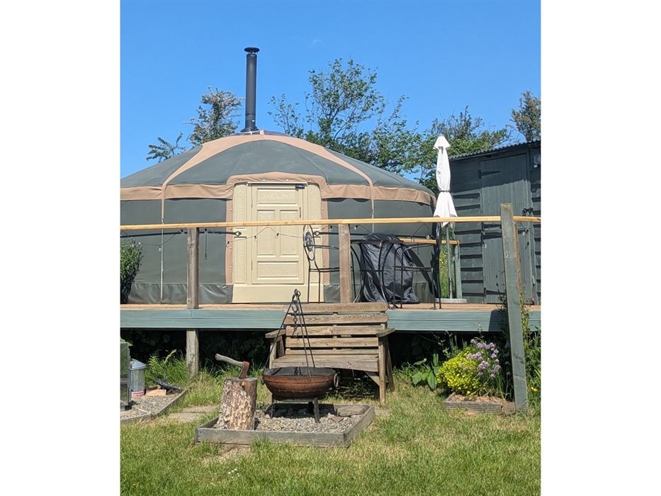 In front of the deck at the yurt, wood bench with wood framed gravel square containing Kadai fire pit with grill and cast iron skillet on the tripod a