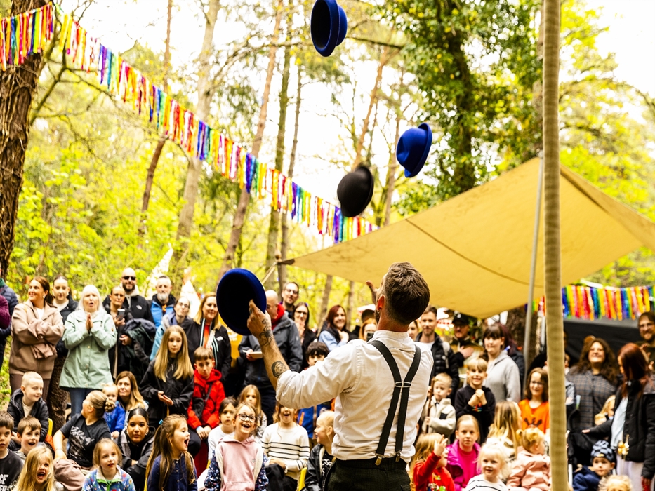 A circus performer juggles hats to an excited audience of families among the trees at Forest Feastival