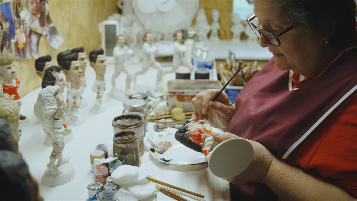 A lady paints a Grogg with red paint, more unfinished Groggs sit on the table.