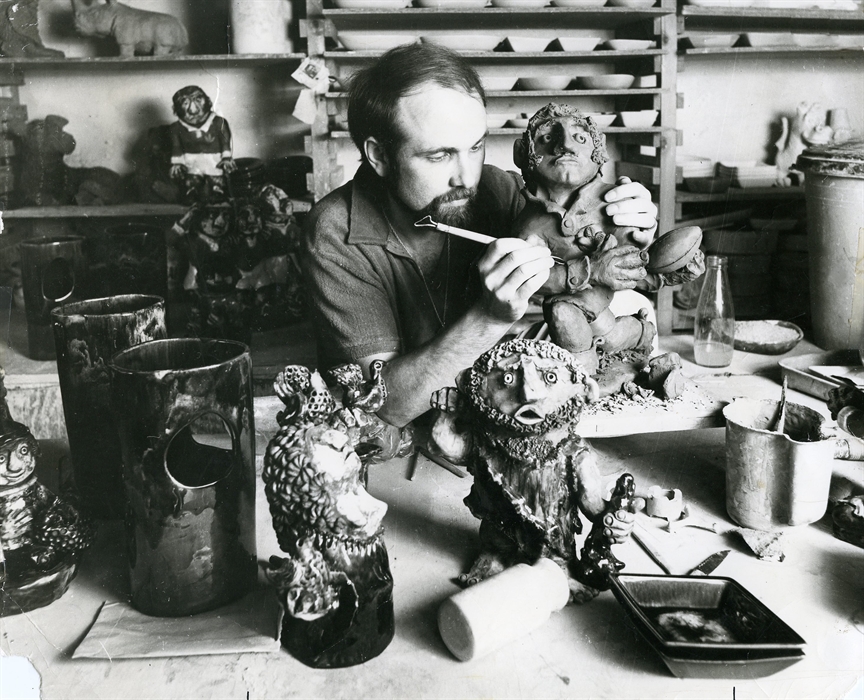 The black and white photo shows John Hughes sculpting a clay rugby Grogg, surrounded by various miscellaneous characters and pottery.