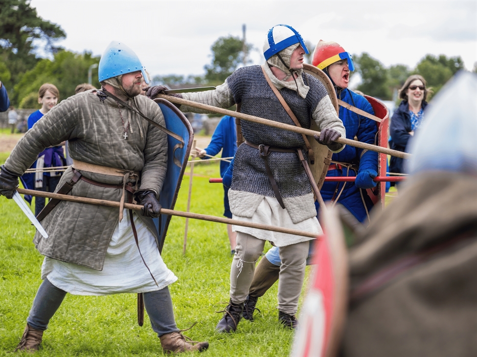 Reenactment of a medieval battle with men in chainmail and helmets holding shields and spears on a grassy field.