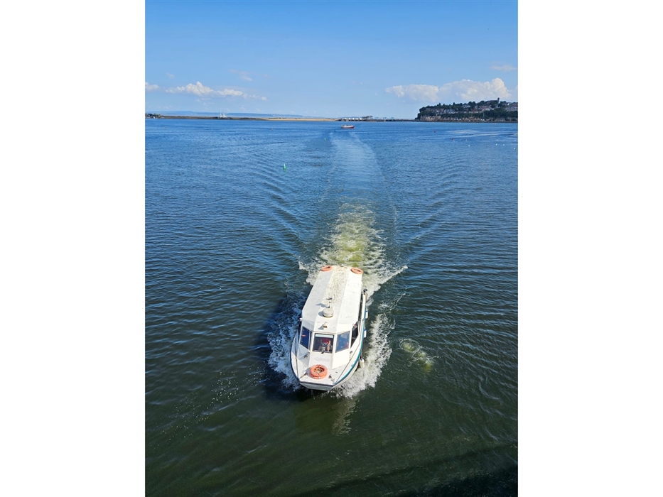 A white Aquabus passenger ferry boat cruising through the blue water of Cardiff Bay, leaving a wake behind it, with the coastline visible in the dista