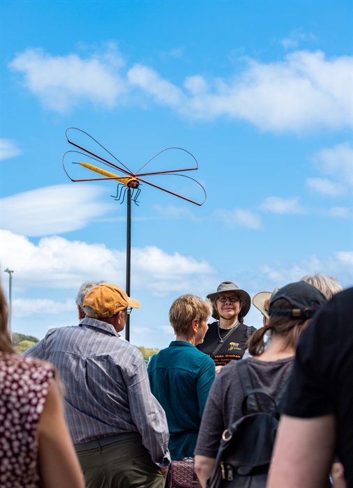 A picture of a tour guide leading a tour group next to a sculpture of a giant dragonfly.