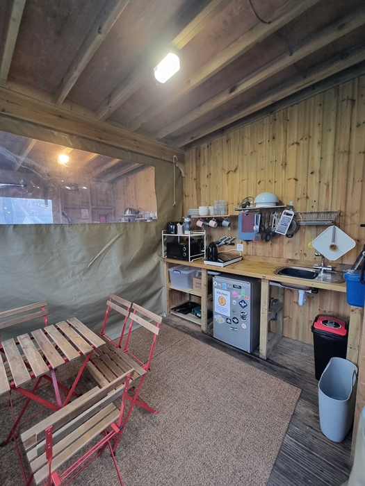 A wooden room with table and chairs and kitchen equipment including a fridge and microwave