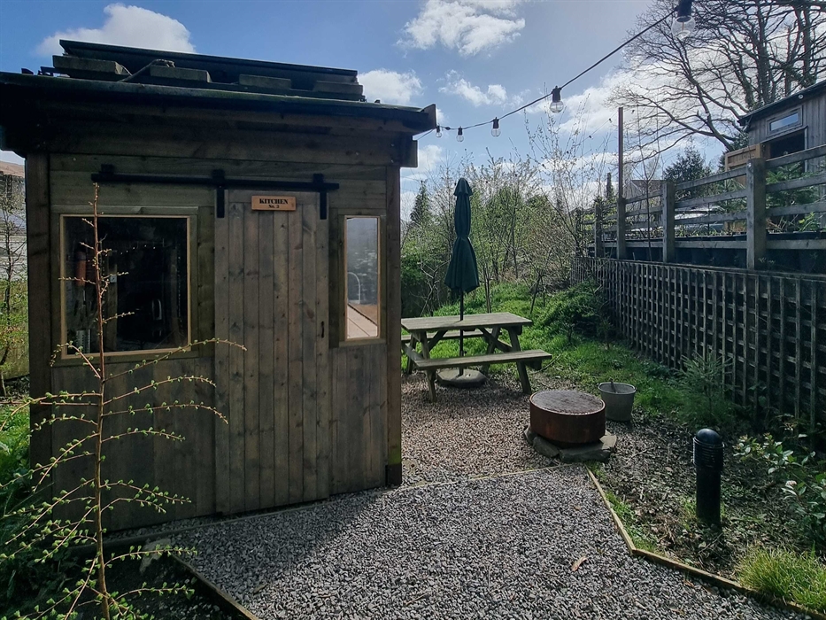 A cabin with a picnic table and firepit surrounded by greenery and a blue cloudy sky